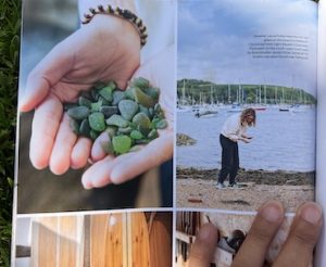 A close up image of National Geographic Traveller Magazine, page 115 showing Laura Polleys hands holding seaglass and her collecting seaglass on the waters edge with boats behind.